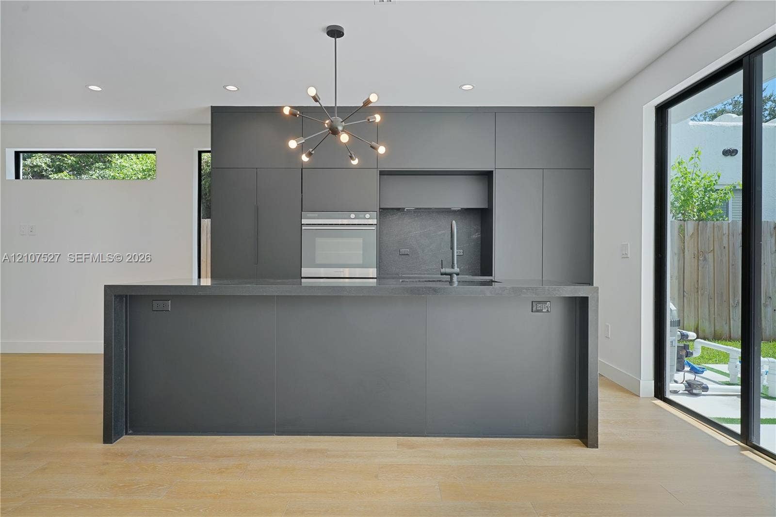 355 NW 50th Street, Miami, FL 33127 Kitchen featuring matte charcoal flat-panel cabinetry, a central island with a waterfall countertop, integrated stainless steel oven, multi-arm chandelier, and wood-finish flooring