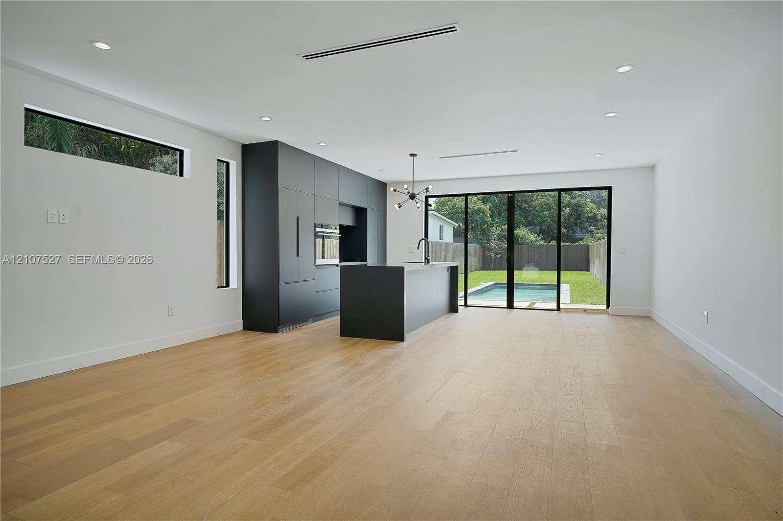 355 NW 50th Street, Miami, FL 33127 Modern kitchen featuring matte charcoal cabinetry, a central island, wood-finish flooring, recessed lighting, and multi-panel glass sliding doors opening to the exterior pool area