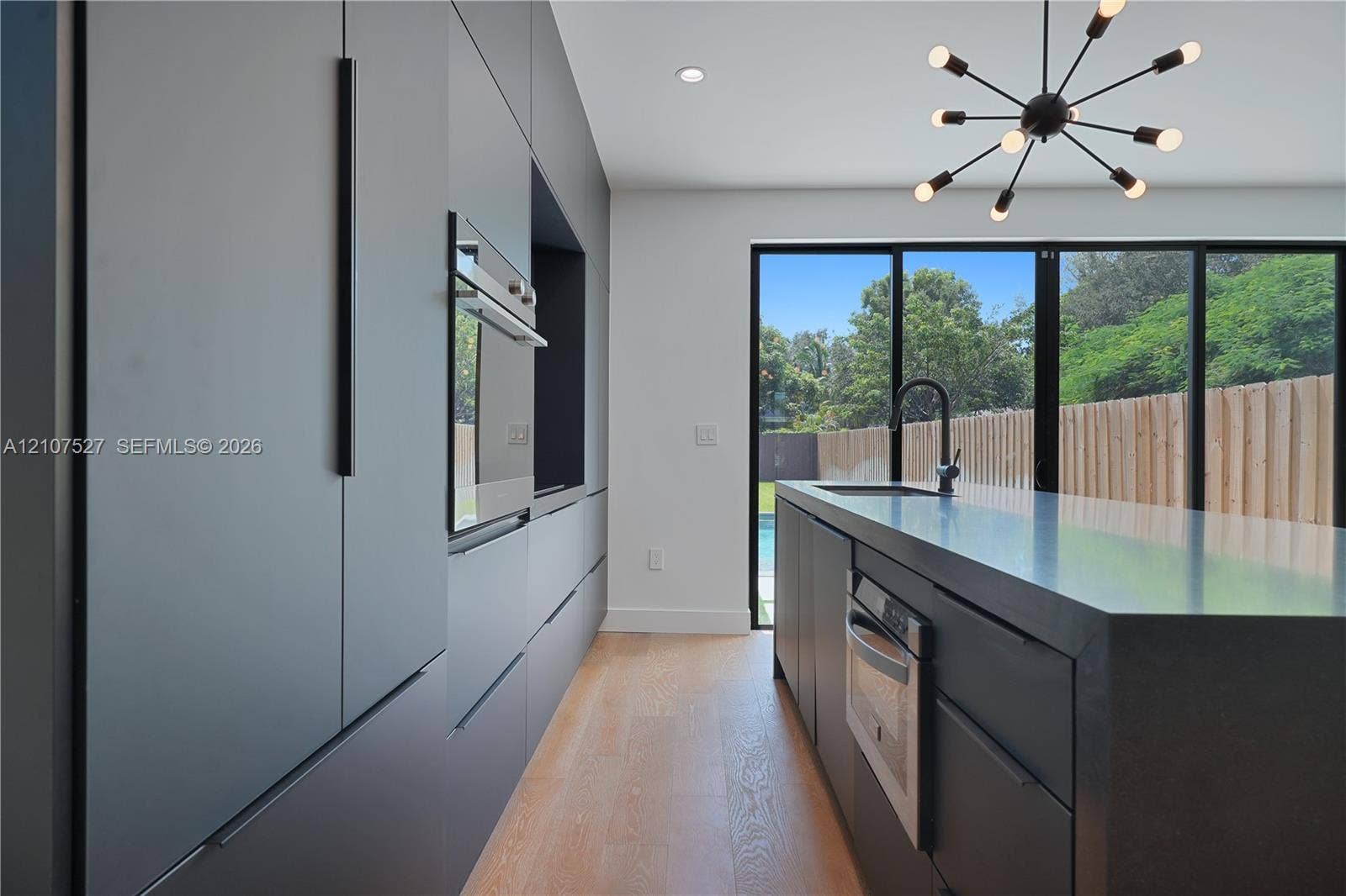 355 NW 50th Street, Miami, FL 33127 Modern kitchen featuring charcoal slab-front cabinetry, an expansive island with a matte black faucet, integrated stainless steel appliances, wood-finish flooring, and multi-panel glass doors