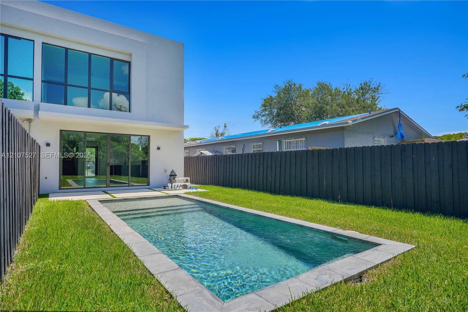 355 NW 50th Street, Miami, FL 33127 Modern exterior featuring multi-pane glass walls, rectilinear in-ground swimming pool with stone coping, manicured lawn, and perimeter wood privacy fencing