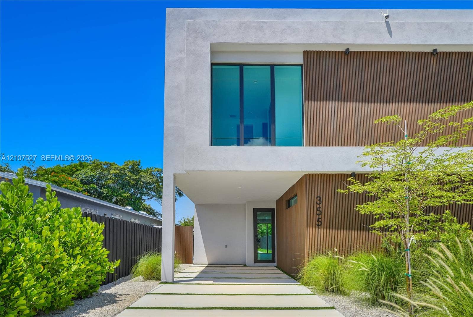 355 NW 50th Street, Miami, FL 33127 Contemporary architectural exterior featuring smooth stucco finish, vertical wood-panel siding, expansive floor-to-ceiling glass windows, and a structured concrete paver driveway with integrated ground cover