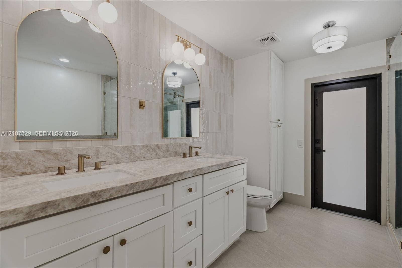 Double vanity with a stone countertop, arched mirrors, brass-finish fixtures, and floor-to-ceiling ceramic tile wall detailing