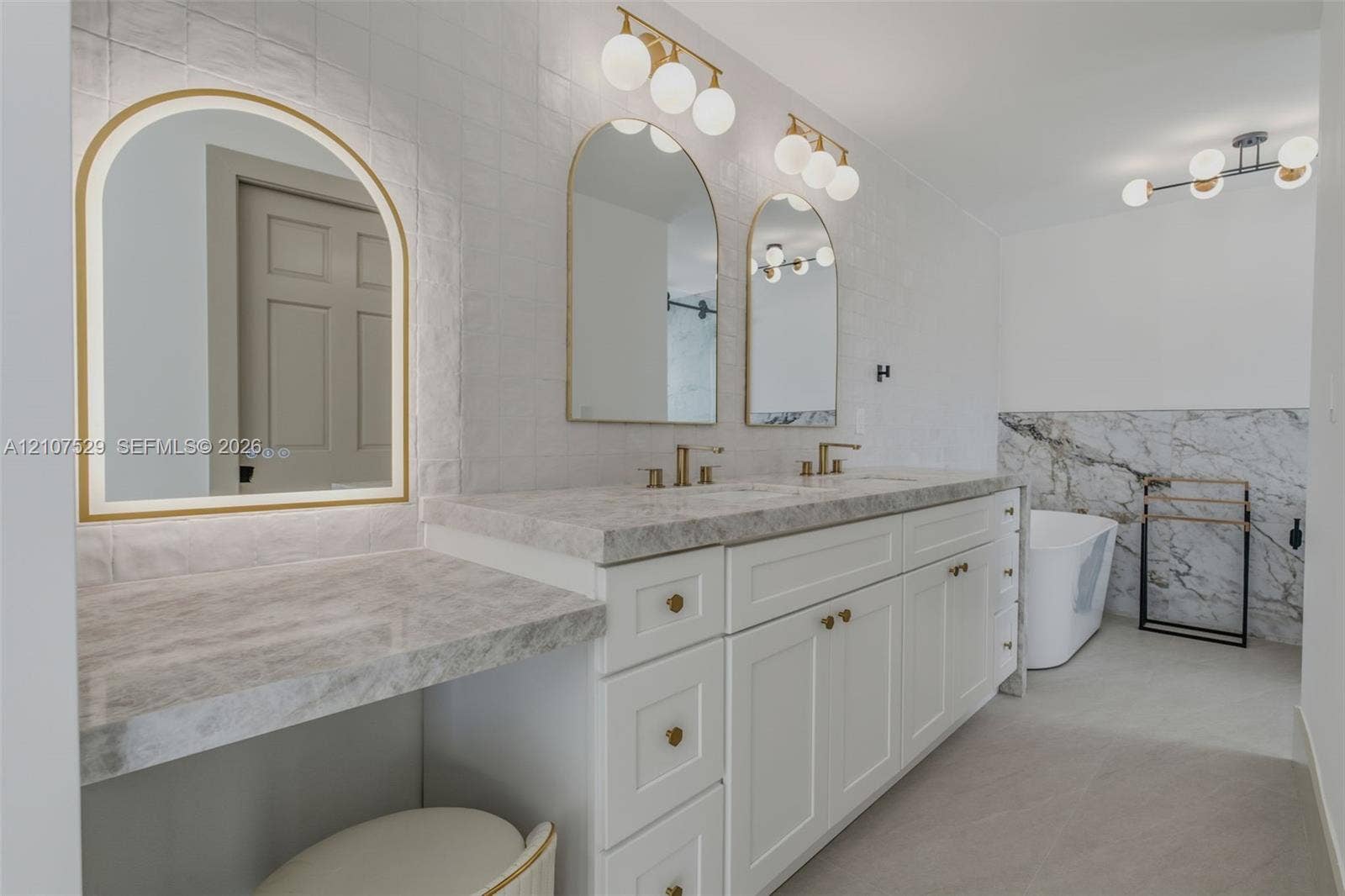 Dual-vanity cabinetry with stone-finish countertops, arched vanity mirrors, multi-bulb lighting, wall-mounted matte black faucet, and a freestanding soaking tub