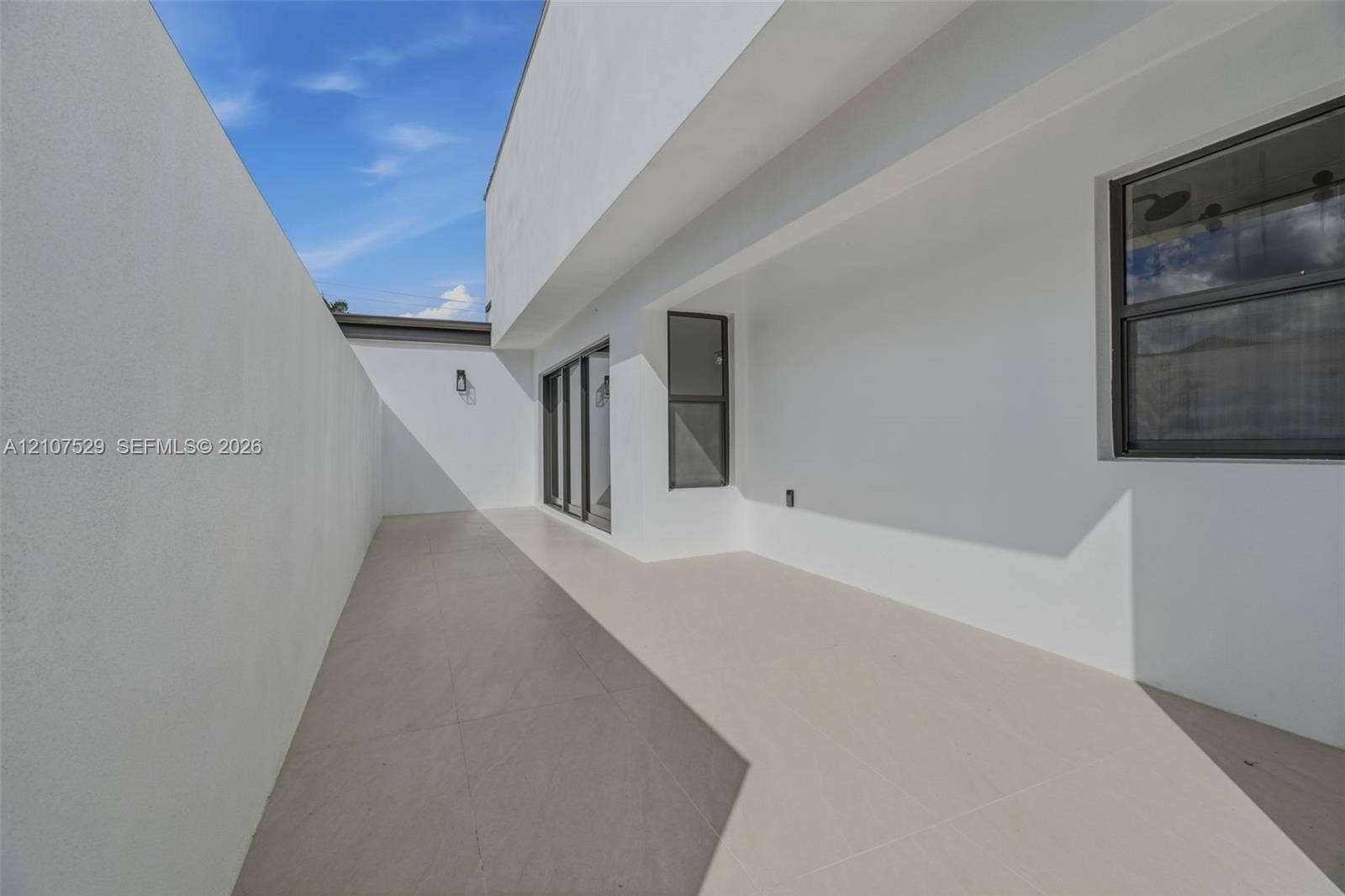 Modern exterior patio featuring large-format porcelain tile flooring, expansive floor-to-ceiling sliding glass panels, smooth white stucco finish, and a private enclosed yard