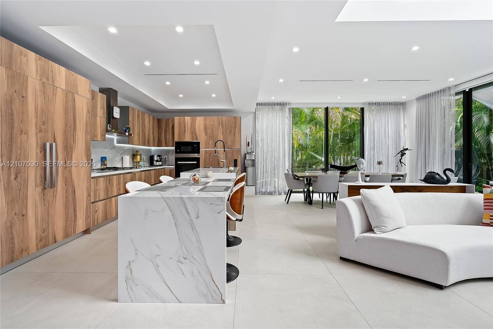 Kitchen featuring wood-finish cabinetry, stone-surfaced waterfall island, large-format tile flooring, recessed ceiling lighting, and floor-to-ceiling glass paneling