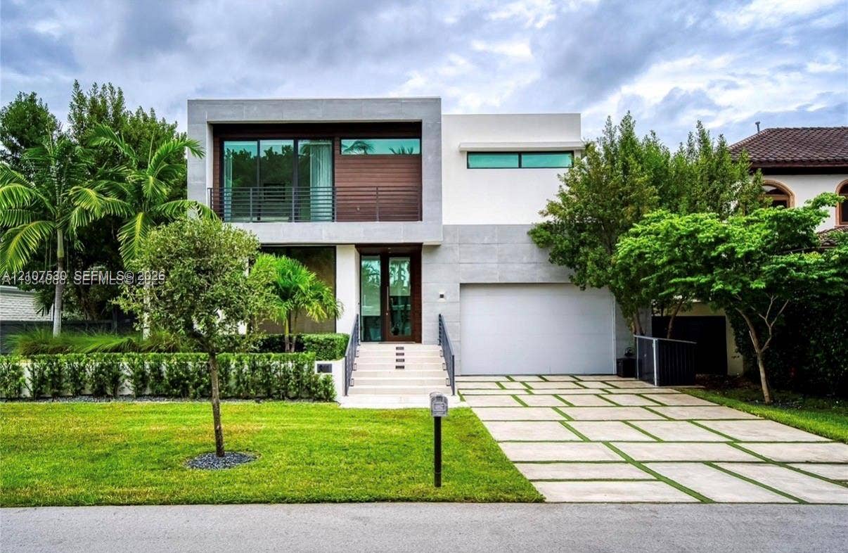 Modern architectural exterior featuring smooth stucco siding, wood-accented panels, a second-story balcony with horizontal metal railing, and a geometric concrete paver driveway