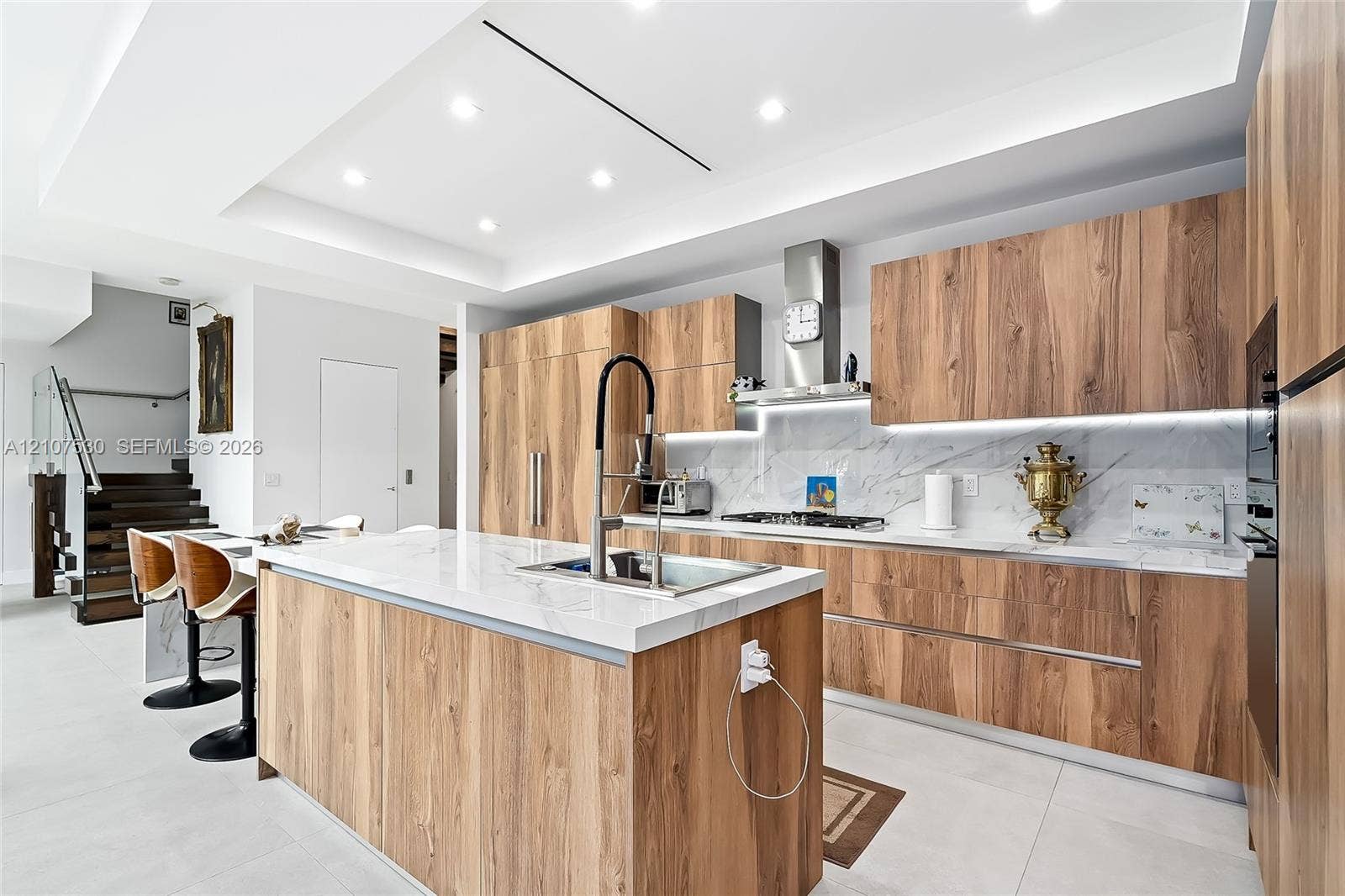 Contemporary kitchen featuring wood-finish cabinetry, stone-patterned countertops, integrated stainless steel appliances, a center island with a high-arch faucet, and recessed ceiling lighting