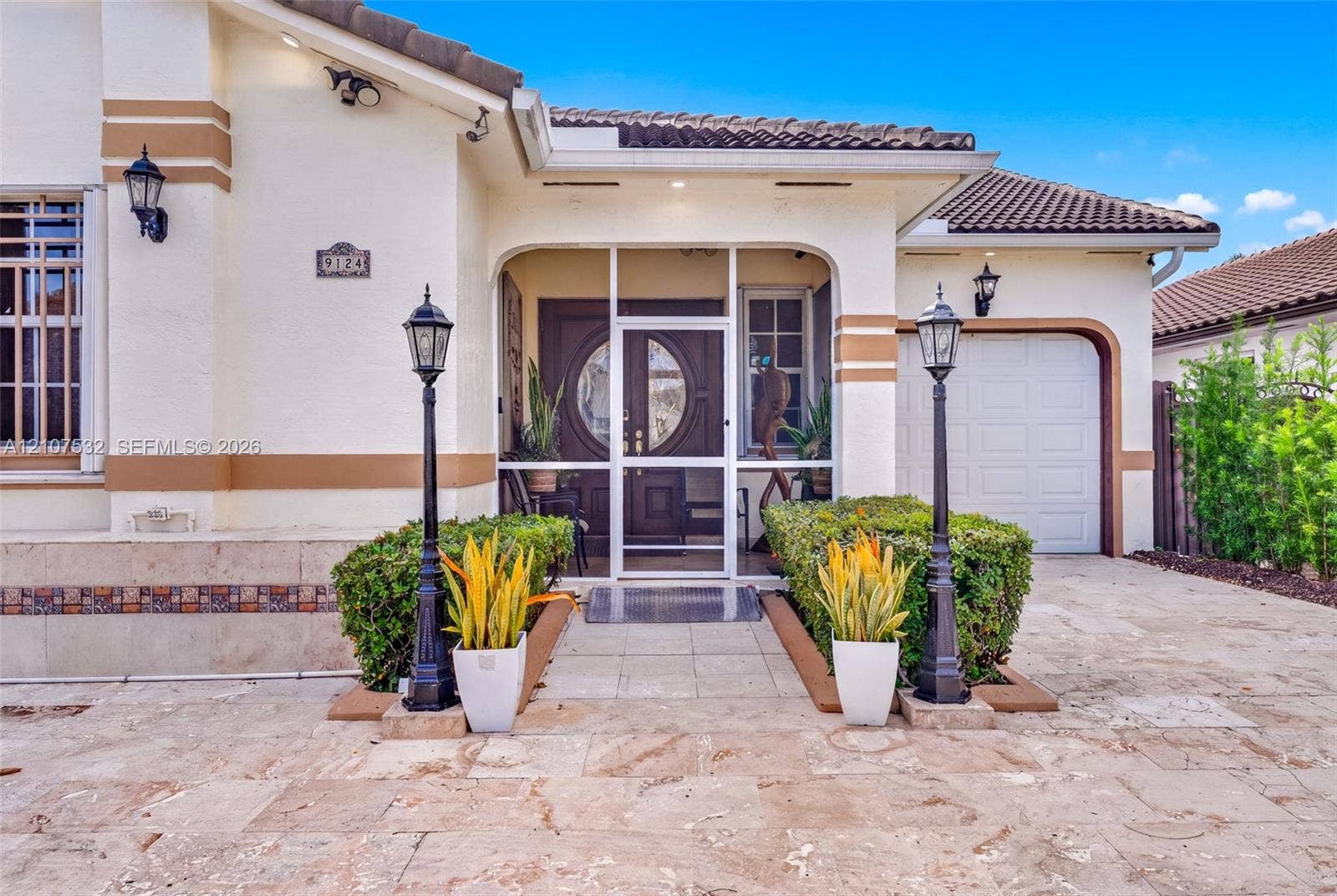 9124 NW 147th Terrace, Miami Lakes, FL 33018 Stucco exterior featuring a tiled roof, arched entryway with a screened enclosure, ornamental iron lamp posts, and stone-finish paved driveway
