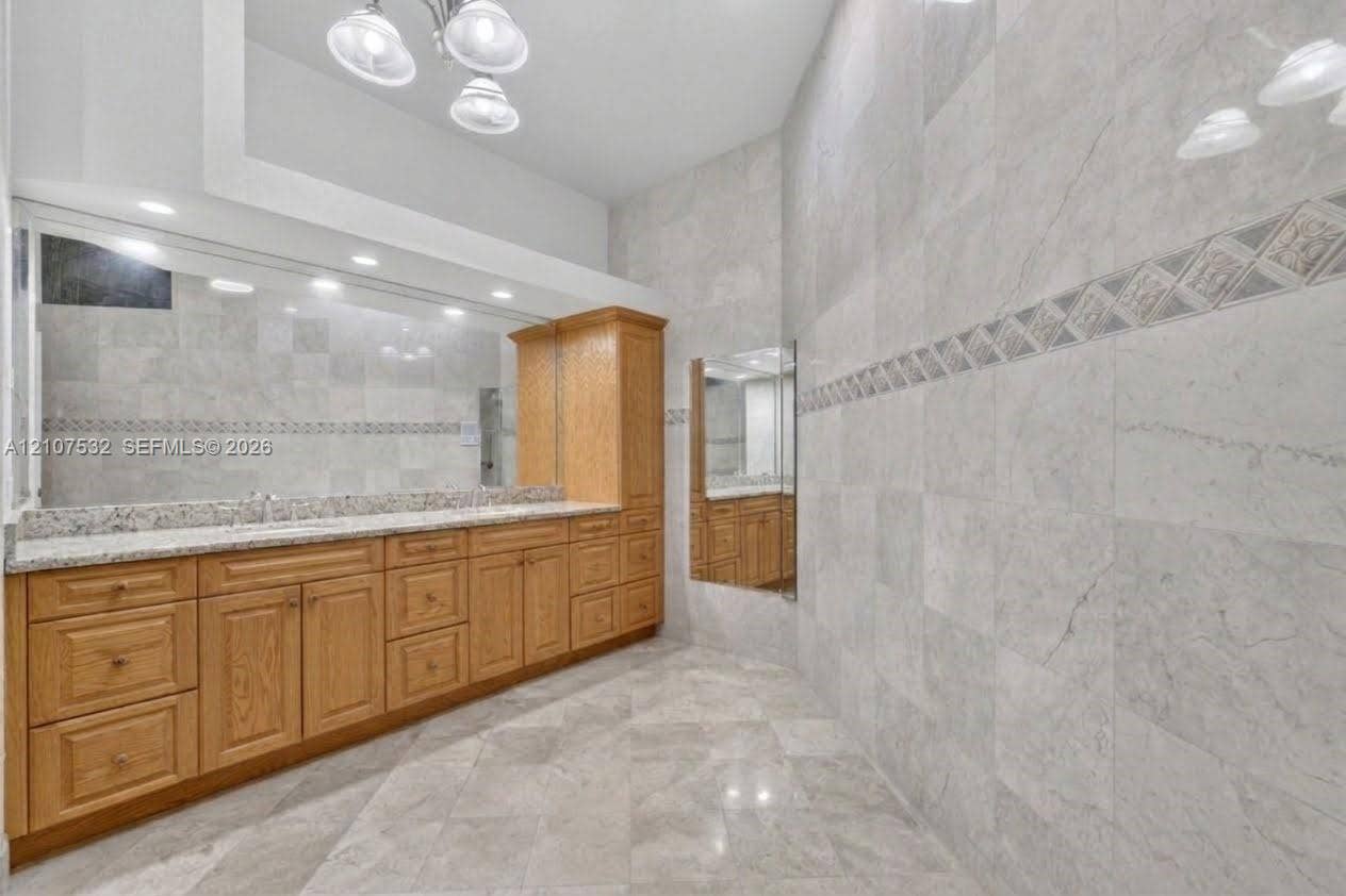 9124 NW 147th Terrace, Miami Lakes, FL 33018 Vanity featuring light-toned wood cabinetry, stone countertop, dual undermount sinks, and full-width mirror, complemented by floor-to-ceiling wall tiling with decorative tile inlay and large-format tile flooring