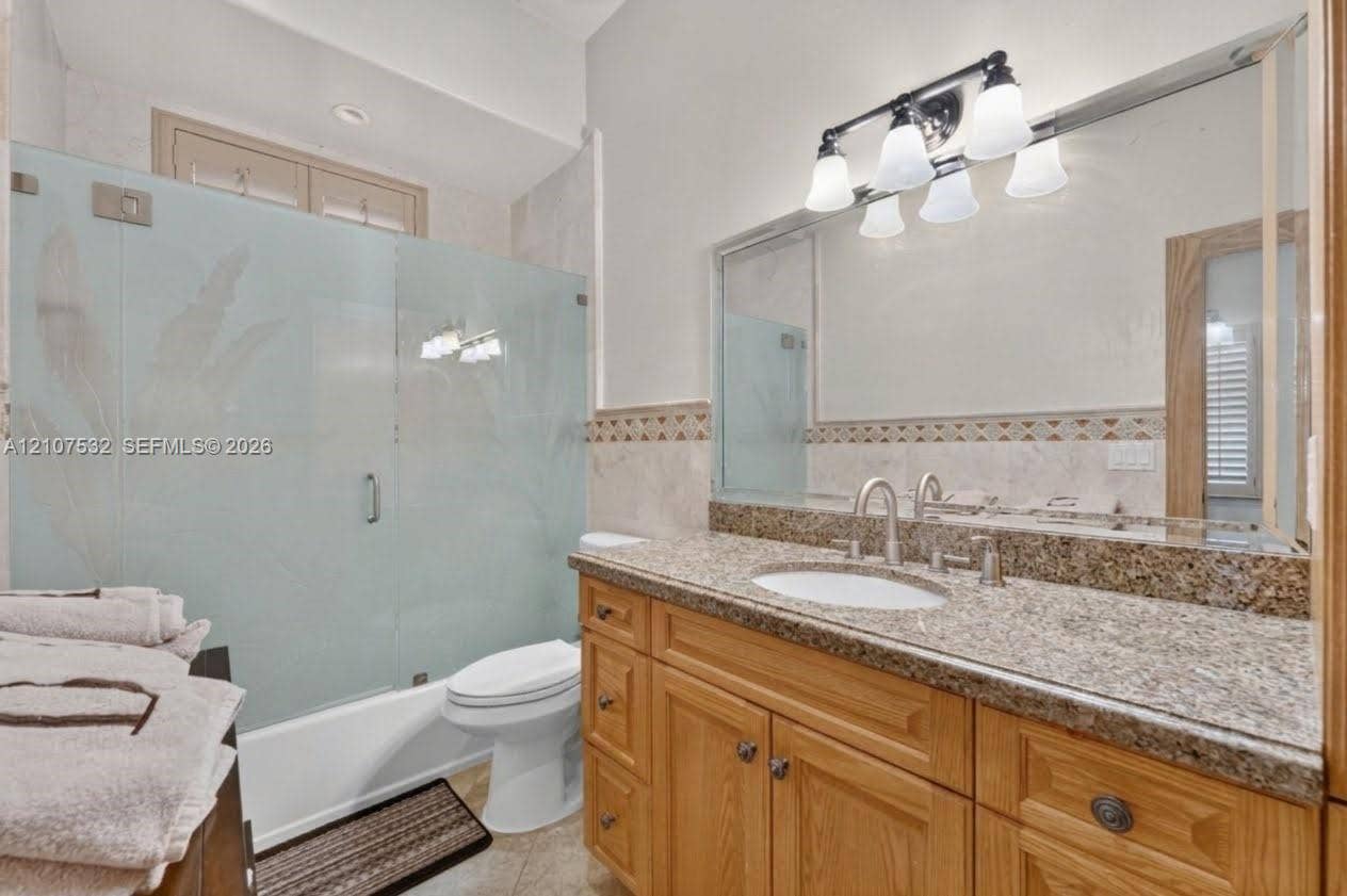 9124 NW 147th Terrace, Miami Lakes, FL 33018 Vanity featuring wood cabinetry, stone-finish countertop, and undermount sink, complemented by a large wall-mounted mirror, four-light vanity fixture, and a tub-shower enclosure with frosted glass paneling
