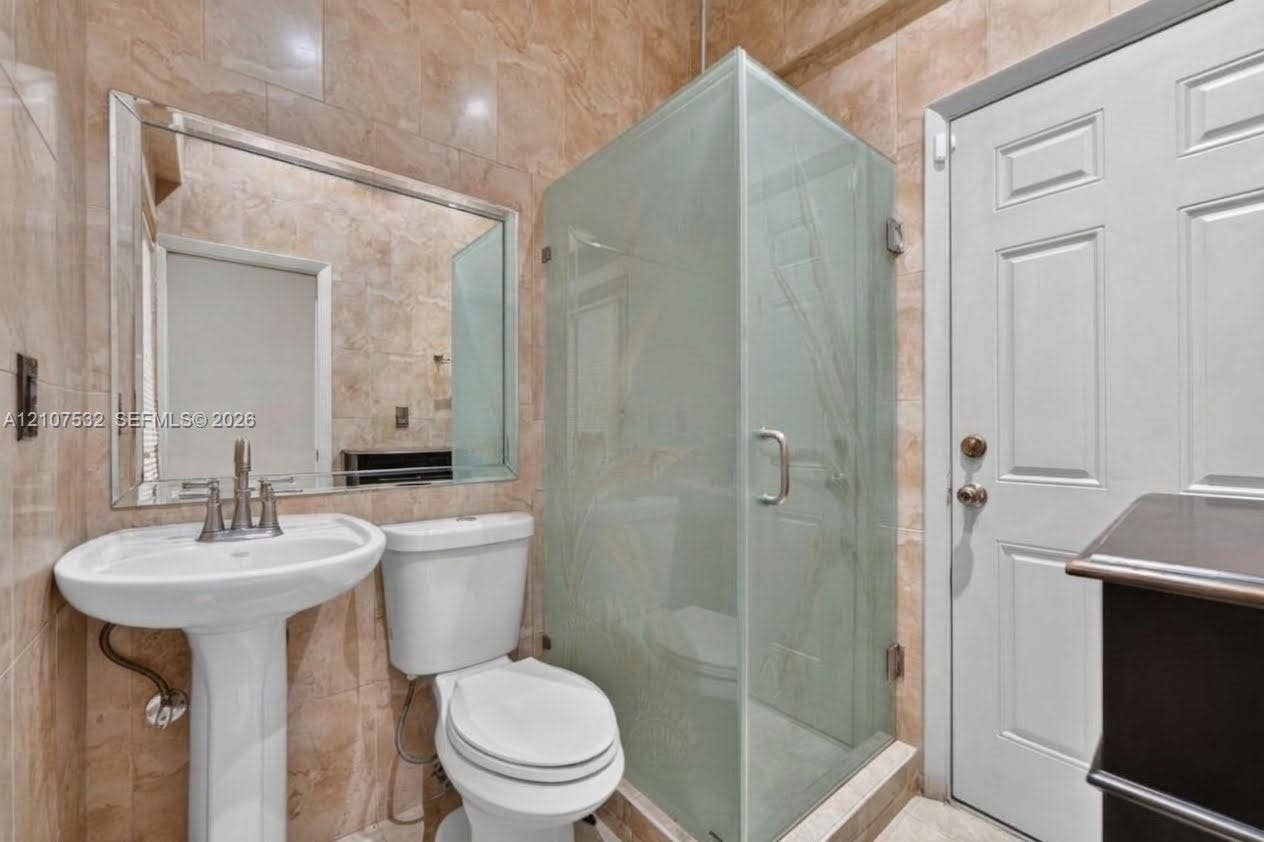 9124 NW 147th Terrace, Miami Lakes, FL 33018 Full-height stone wall tiling, a pedestal sink with a dual-handle faucet, a framed wall mirror, a glass-enclosed shower with etched detail, and a raised-panel door
