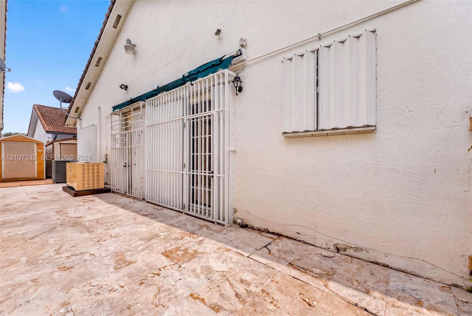 9124 NW 147th Terrace, Miami Lakes, FL 33018 Exterior area featuring a neutral-toned stone-tile patio, white stucco siding, metal security shutters, and an attached retractable awning