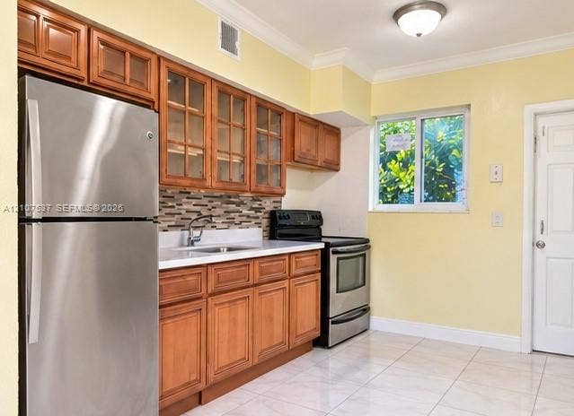 Kitchen featuring wood cabinetry with glass-front uppers, stainless steel appliances, multi-toned tile backsplash, solid-surface countertops, and tile flooring