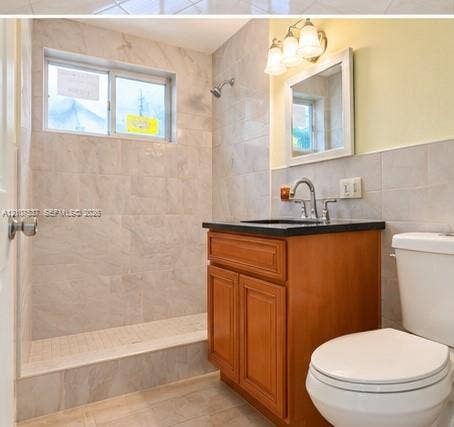 Bathroom featuring a wood-finish vanity with a dark stone-top surface, wall-mounted three-light fixture, framed mirror, and tile-surround walk-in shower