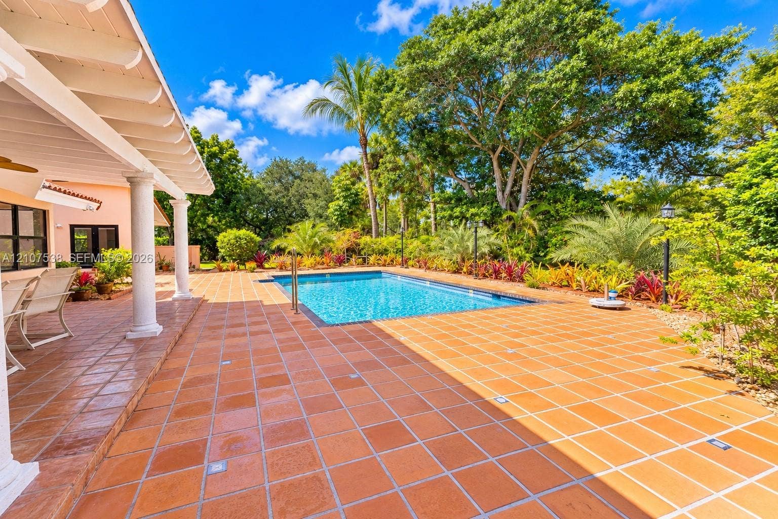 10204 SW 96th Court, Miami, Miami, FL 33176 Covered lanai with white structural columns, expansive terracotta-tiled patio, rectangular swimming pool, and lush perimeter landscaping featuring mature trees and assorted tropical vegetation