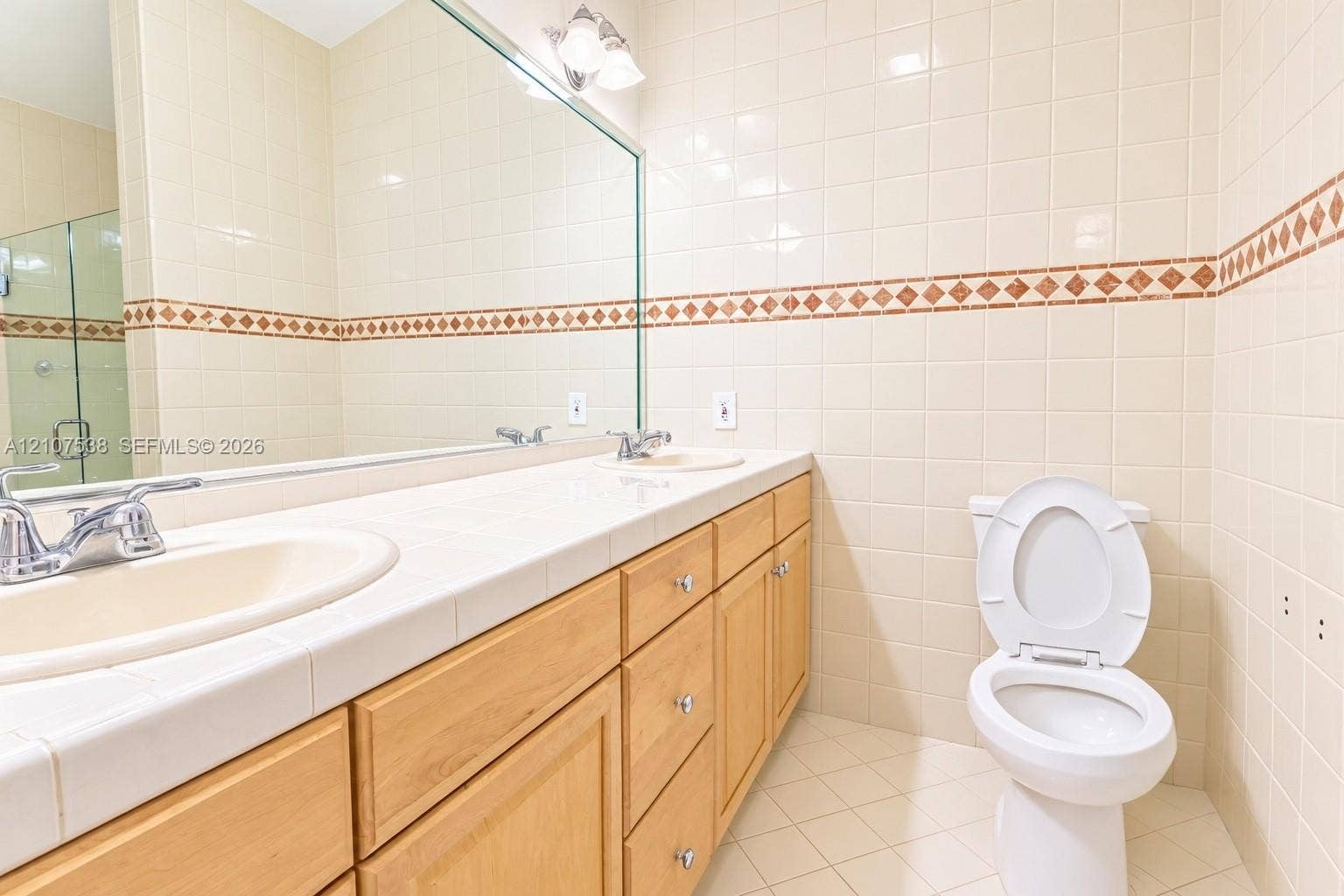 10204 SW 96th Court, Miami, Miami, FL 33176 Bathroom featuring a dual-sink vanity with a tile countertop, light wood-finish cabinetry, a full-width wall mirror, and floor-to-ceiling tiled walls with decorative mosaic accents