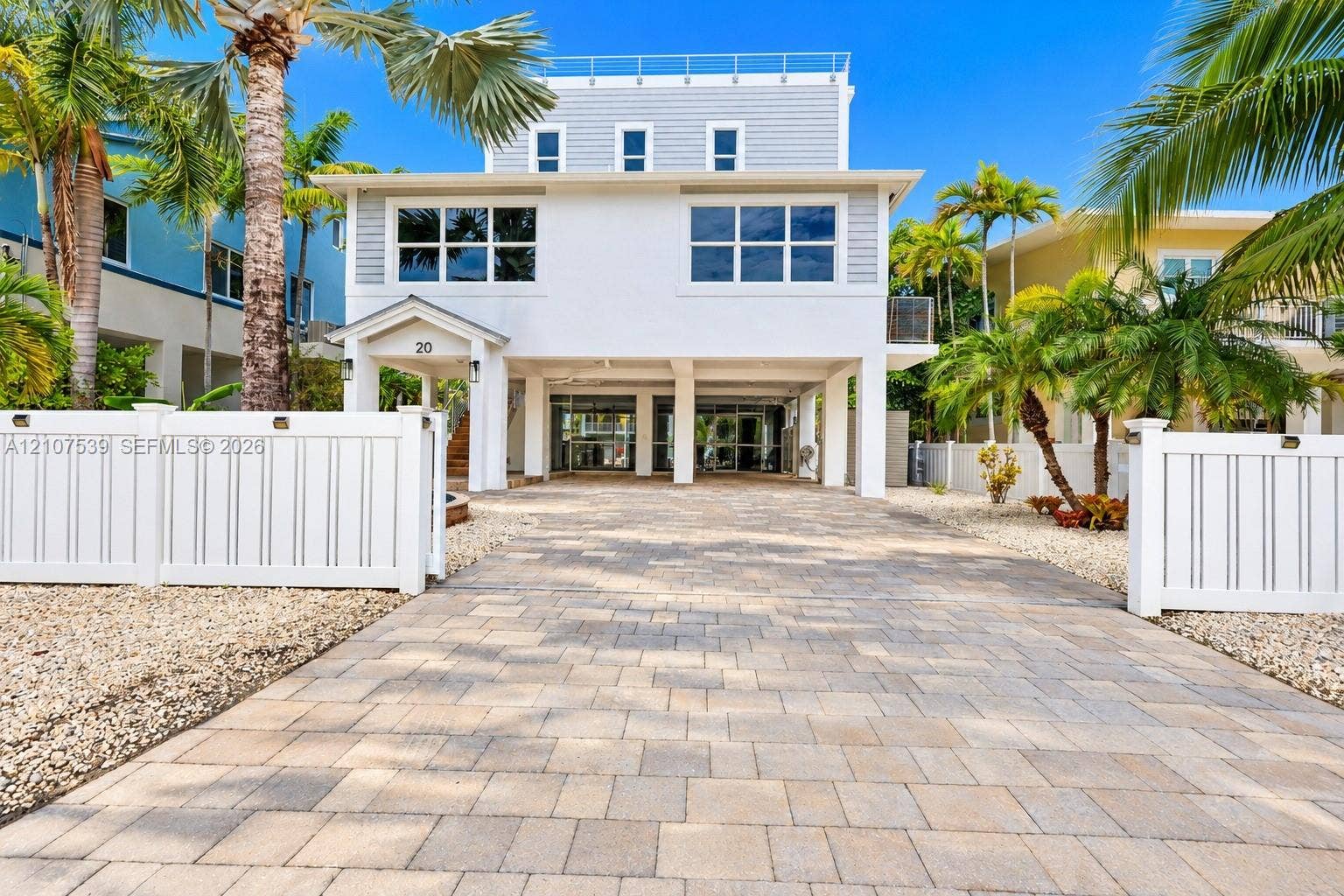20 Center Lane, Key Largo, FL 33037 Three-story elevated residence featuring a paver driveway, covered ground-level parking, multi-pane windows, and a white vinyl perimeter fence