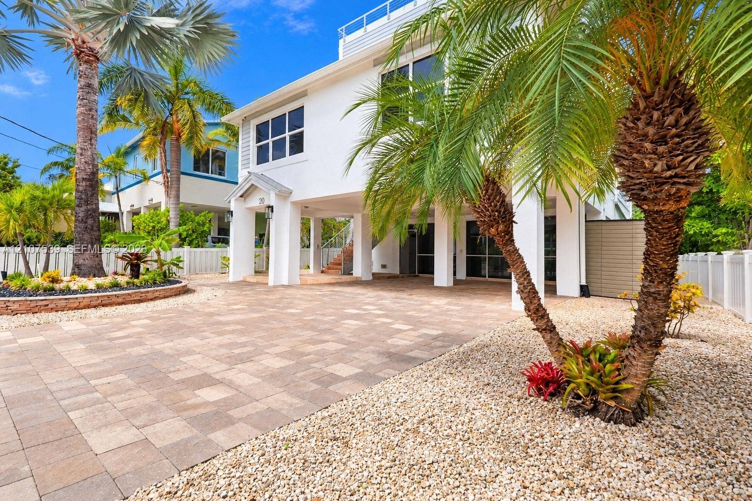 20 Center Lane, Key Largo, FL 33037 Two-story elevated residence featuring a paver driveway, covered ground-level parking, white siding, and landscaped exterior grounds with palm trees