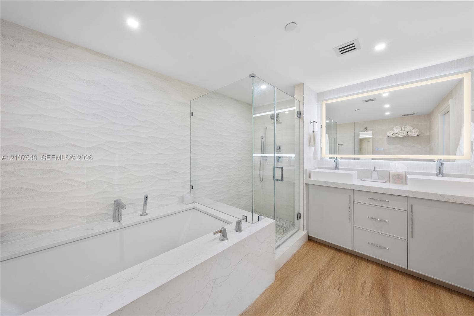 Master bathroom featuring a frameless glass-enclosed shower, a deep soaking tub with stone-finish surround, a dual-sink vanity with comfort height cabinetry, and an oversized back illuminated wall mirror