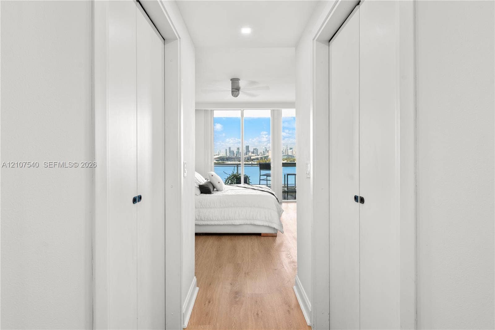 Master bedroom hallway with dual bifold closet doors, wood-finish flooring, recessed LED lighting, and floor-to-ceiling windows with views of the downtown Miami skyine + Biscayne Bay