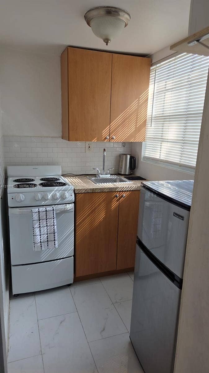 6431 Hayes Street, #3, Hollywood, Hollywood, FL 33024 Kitchenette featuring wood-finish cabinetry, subway tile backsplash, light-toned stone-finish countertops, ceramic tile flooring, and integrated stainless steel and white appliances