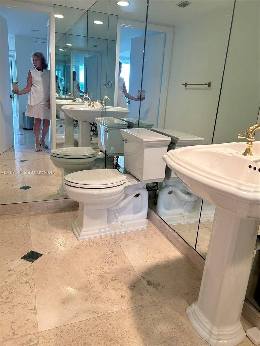 19333 Collins Avenue, #2007, Sunny Isles Beach, Sunny Isles Beach, FL 33160 Bathroom featuring large-format light stone flooring with inlay accents, a pedestal sink with gold-tone hardware, and floor-to-ceiling wall mirrors