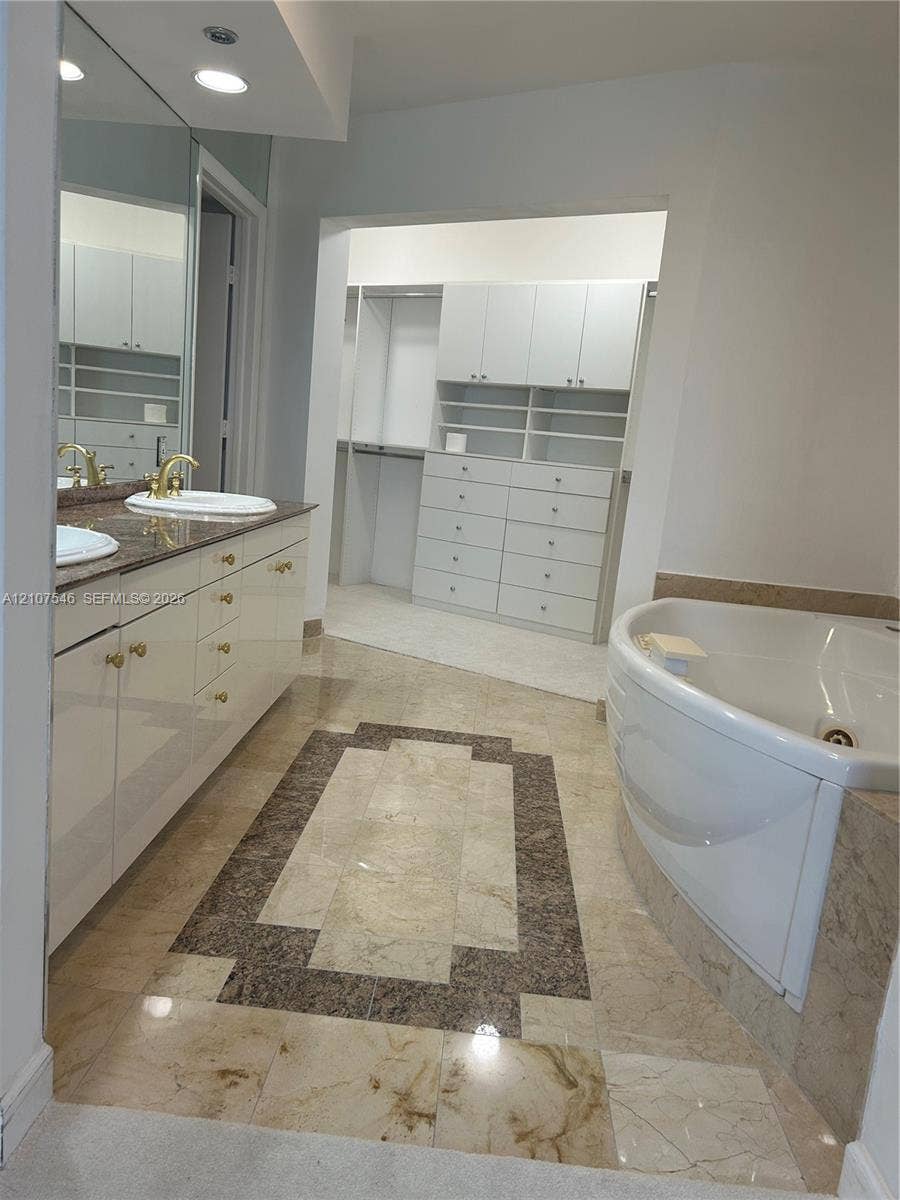 19333 Collins Avenue, #2007, Sunny Isles Beach, Sunny Isles Beach, FL 33160 Dual-sink vanity with stone countertop and gold-tone fixtures, expansive wall mirror, jetted soaking tub with stone surround, and custom built-in wardrobe cabinetry with integrated drawer storage
