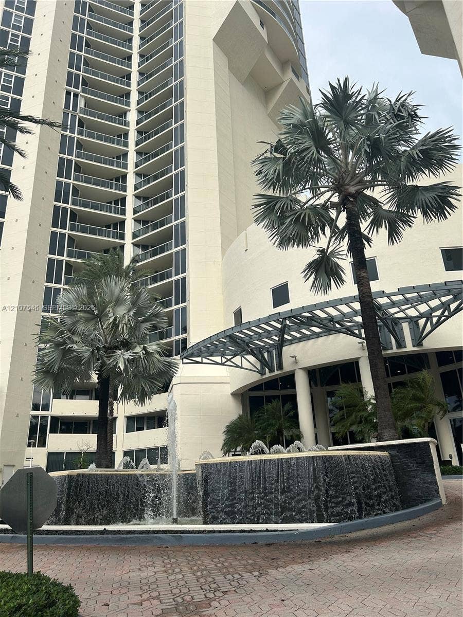 19333 Collins Avenue, #2007, Sunny Isles Beach, Sunny Isles Beach, FL 33160 High-rise exterior featuring curved glass balconies, a circular fountain with tiered stone masonry, a metal-framed porte-cochere, and mature palm landscaping