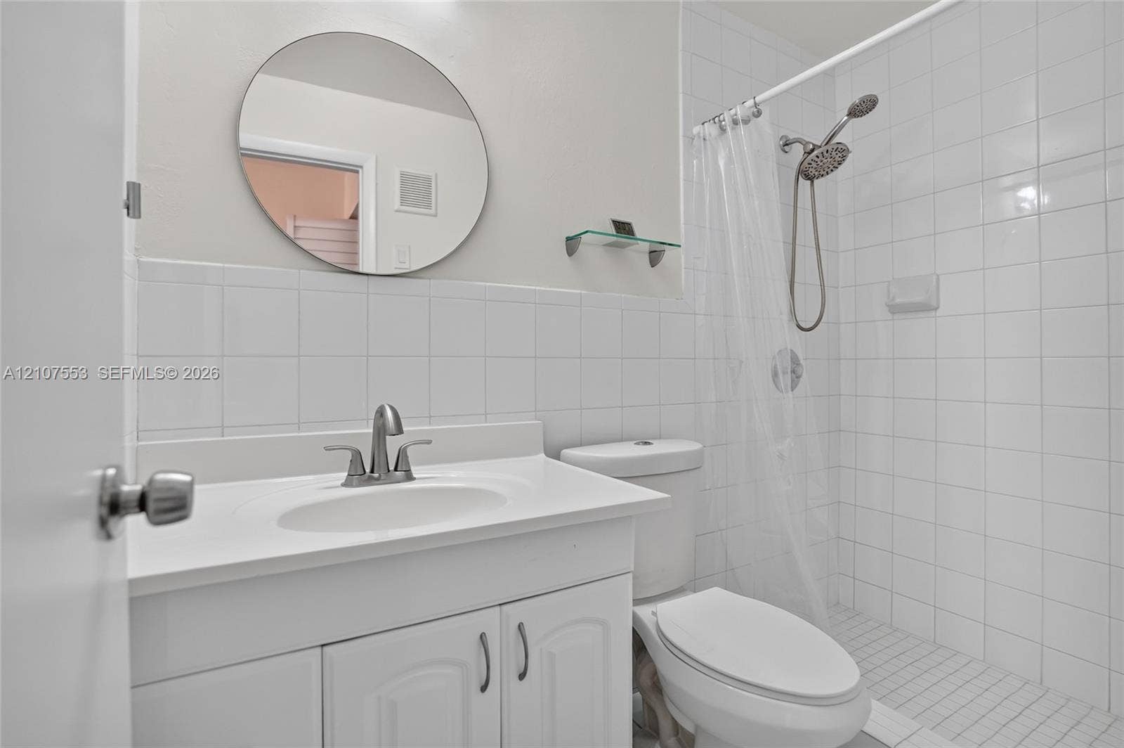 7336 SW 26th Court, #51, Davie, Davie, FL 33314 Bathroom featuring a white vanity cabinet with an integrated basin, circular wall-mounted mirror, glass shelf, and a walk-in shower enclosure with white wall tile