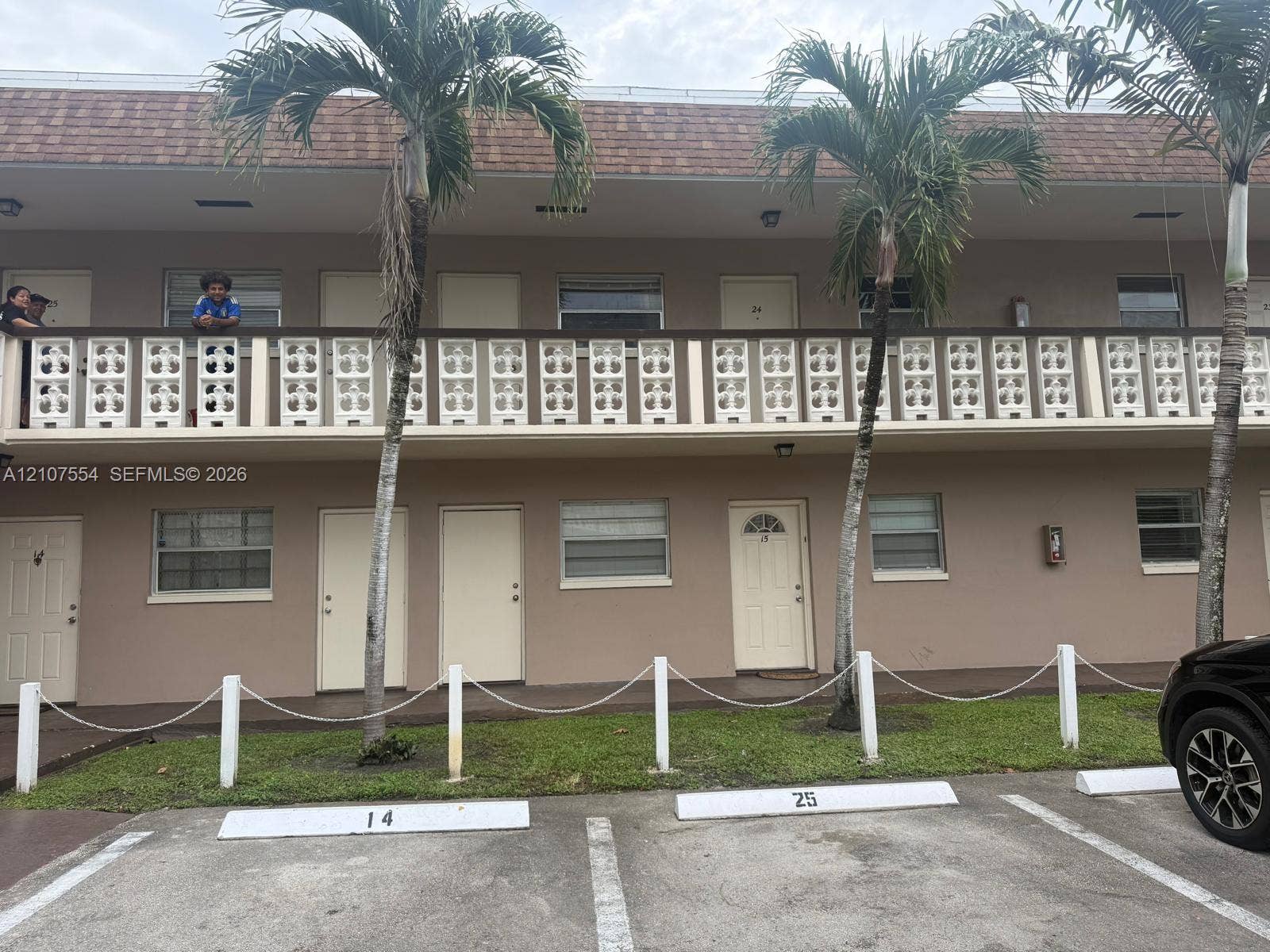 Two-story multi-unit exterior featuring decorative breeze block balcony railings, shingled mansard roofline, and assigned paved parking spaces