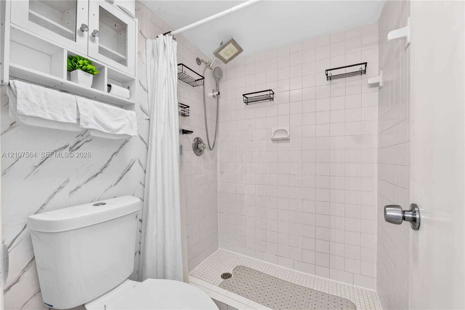Bathroom featuring a shower stall with white tile surround, a built-in overhead showerhead with an adjustable handheld attachment, wall-mounted shelving, and white cabinetry