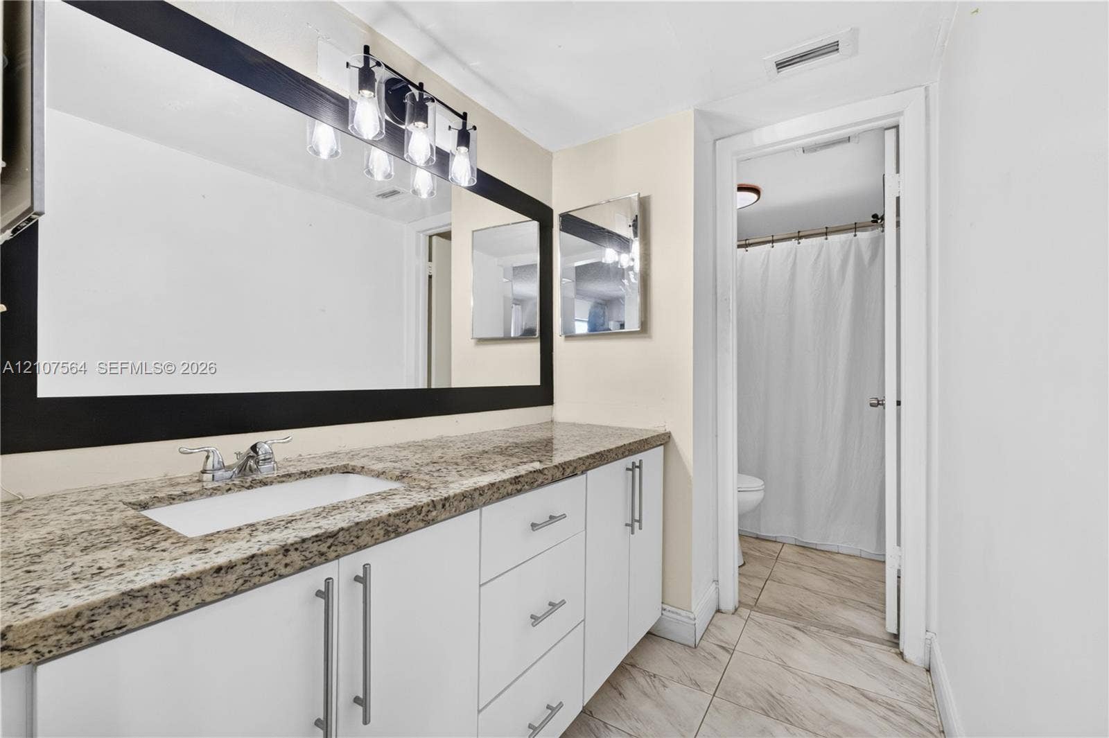 White cabinetry with stainless steel pulls, speckled stone countertop, large rectangular mirror with a dark frame, four-light vanity fixture, and tile flooring