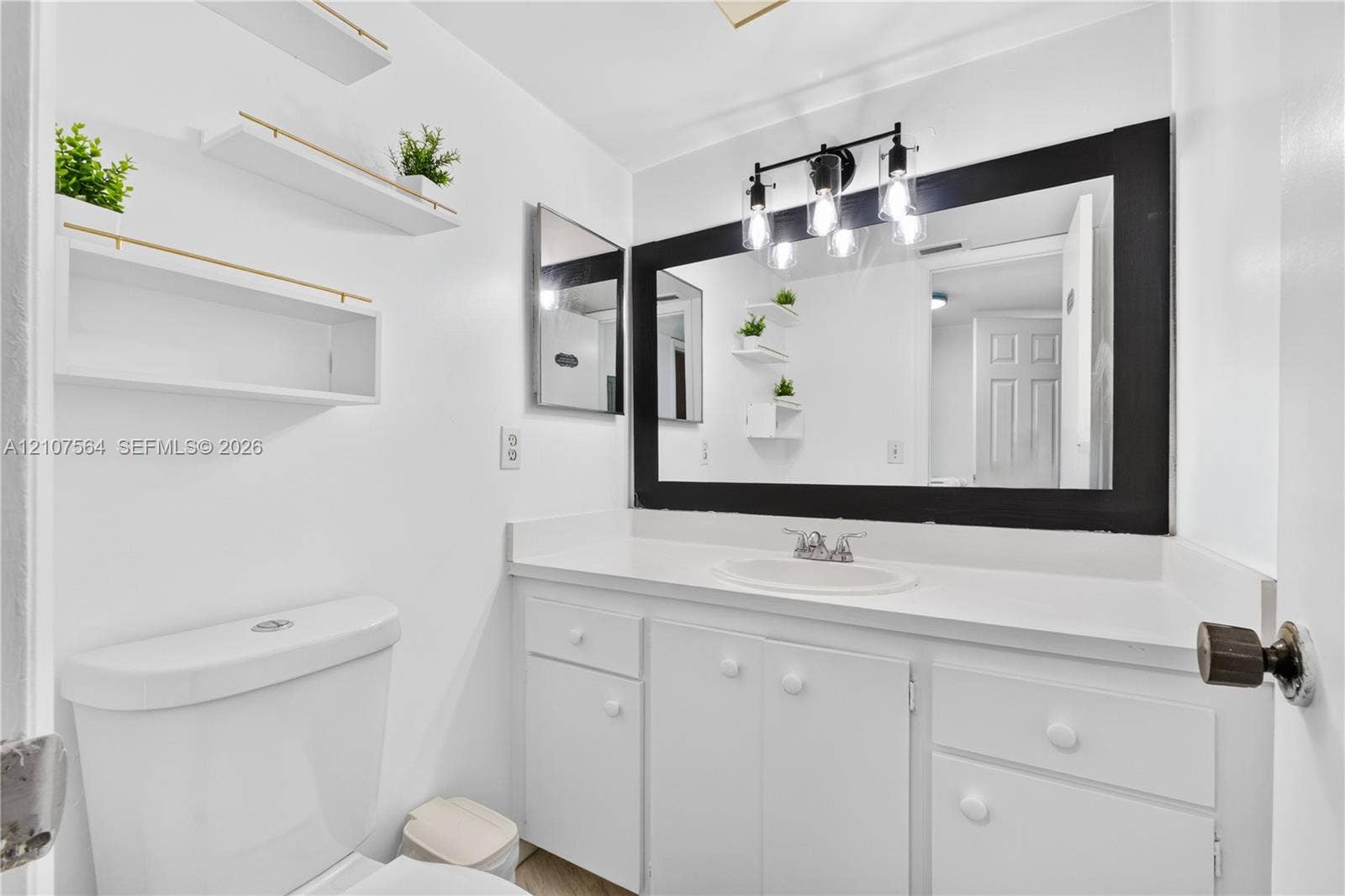 Bathroom featuring a vanity with a white countertop, inset sink, three-light fixture, large black-framed mirror, and wall-mounted white shelving