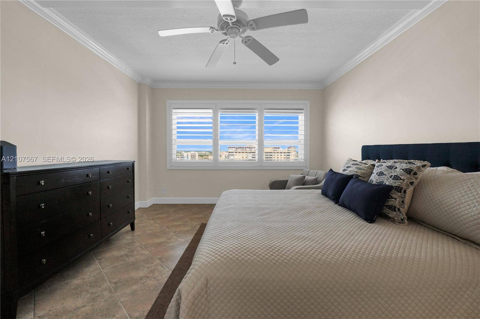 Neutral-toned interior featuring large-format tile flooring, multi-pane window configuration with plantation-style shutters, ceiling-mounted paddle fan, and crown molding