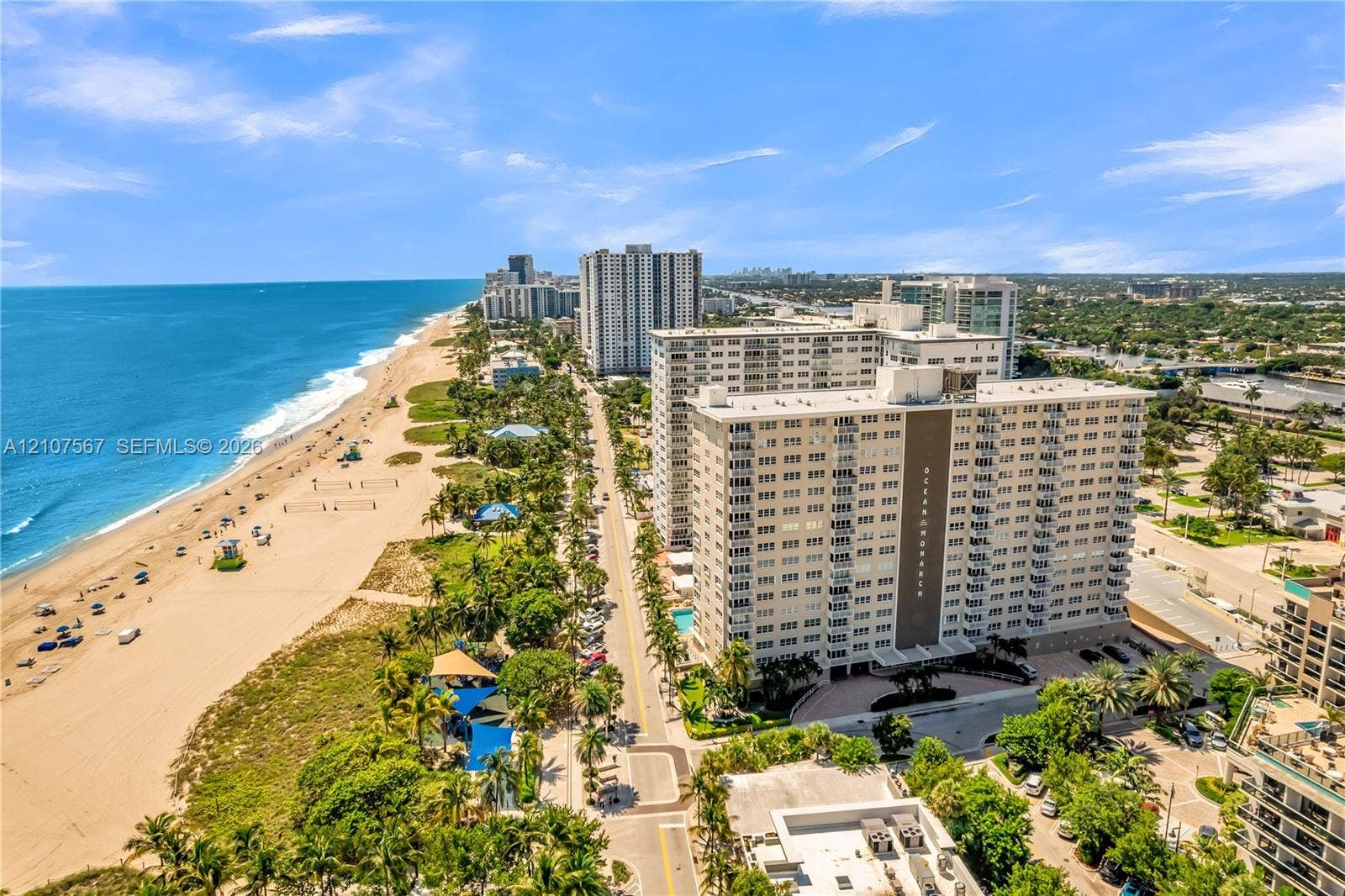 Aerial perspective of coastal high-rise property featuring direct proximity to the ocean, expansive sandy beach, mature palm tree landscaping, and paved parking areas