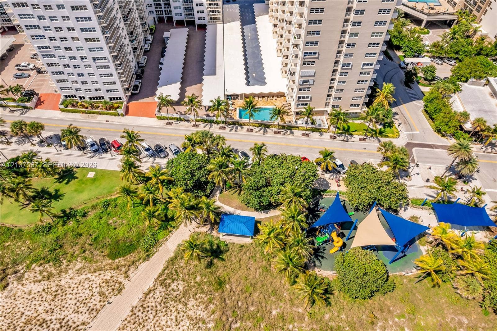 Aerial perspective featuring a high-rise residential structure with covered parking, a swimming pool, and proximity to a coastal park with shaded playground equipment and sandy terrain
