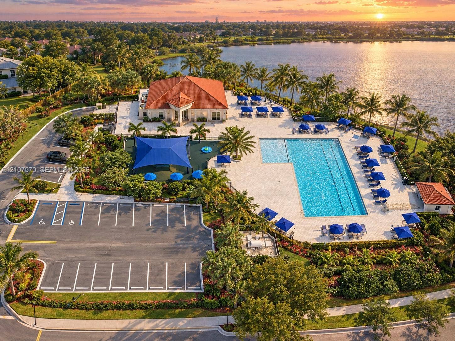 Waterfront community amenity complex featuring a rectangular swimming pool, expansive paved patio, shaded recreational area, and paved parking lot