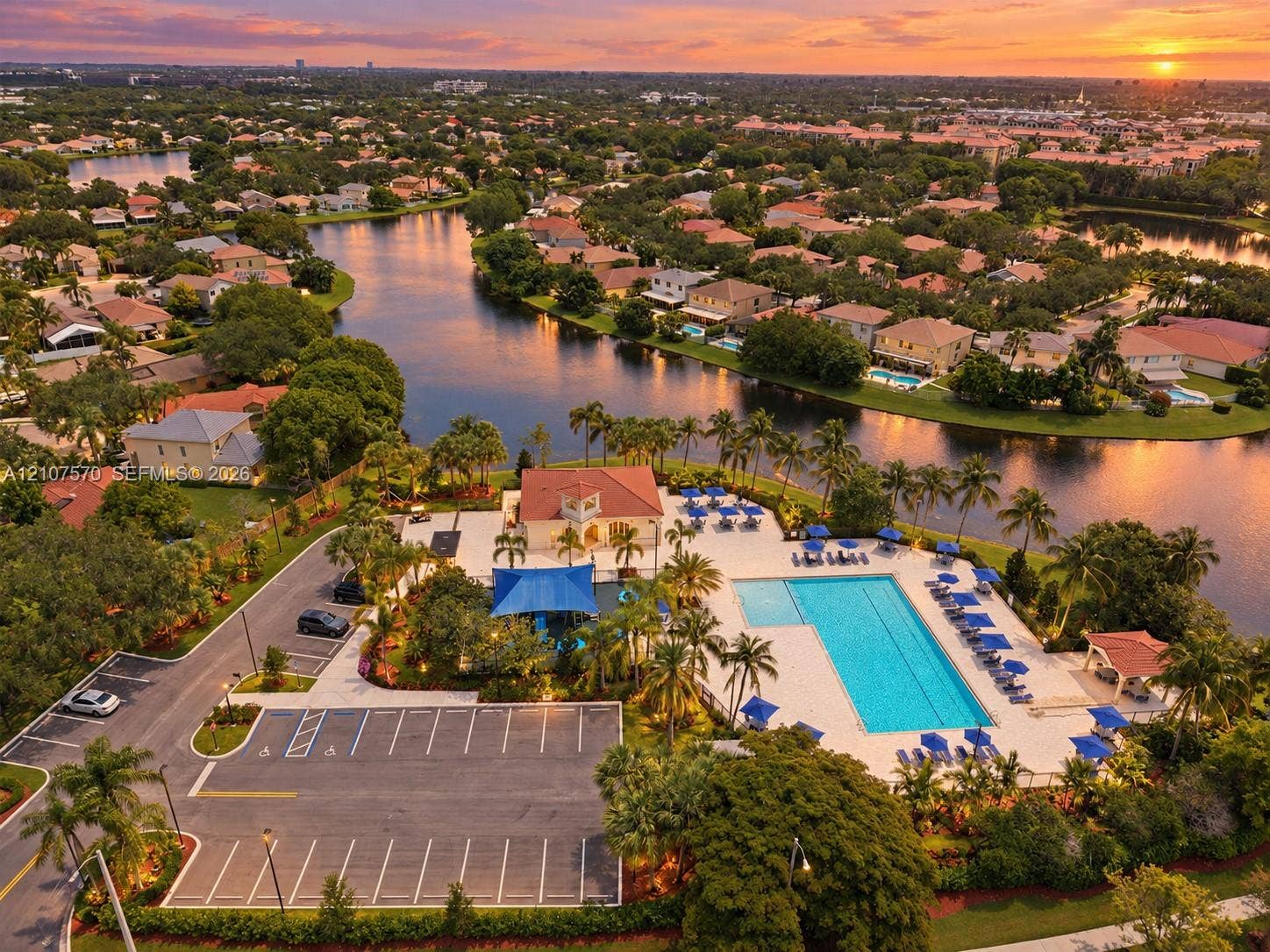 Aerial perspective of the community recreation facility featuring a swimming pool, paved parking area, clubhouse building, and mature palm trees situated along the waterfront