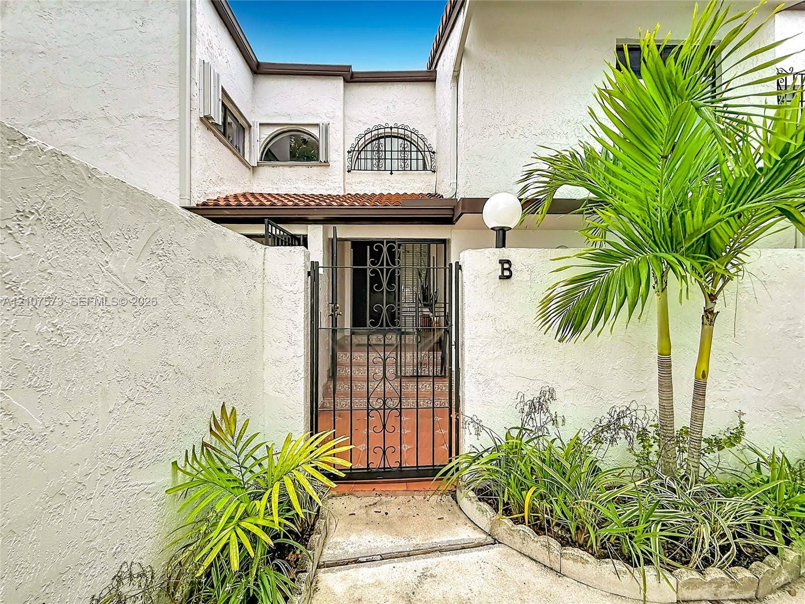 1291 SW 124th Court, #19-B, Miami, FL 33184 Stucco exterior featuring arched windows with decorative metal grilles, a gated courtyard entry, terracotta-style paving, and attached globe lighting