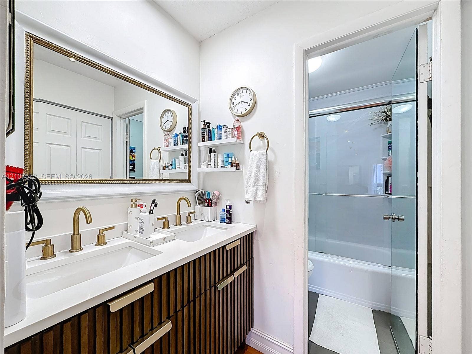 1291 SW 124th Court, #19-B, Miami, FL 33184 Double vanity featuring a white countertop, dual undermount sinks, brass-finish faucets, vertical-slat cabinetry with metallic hardware, and a framed wall mirror