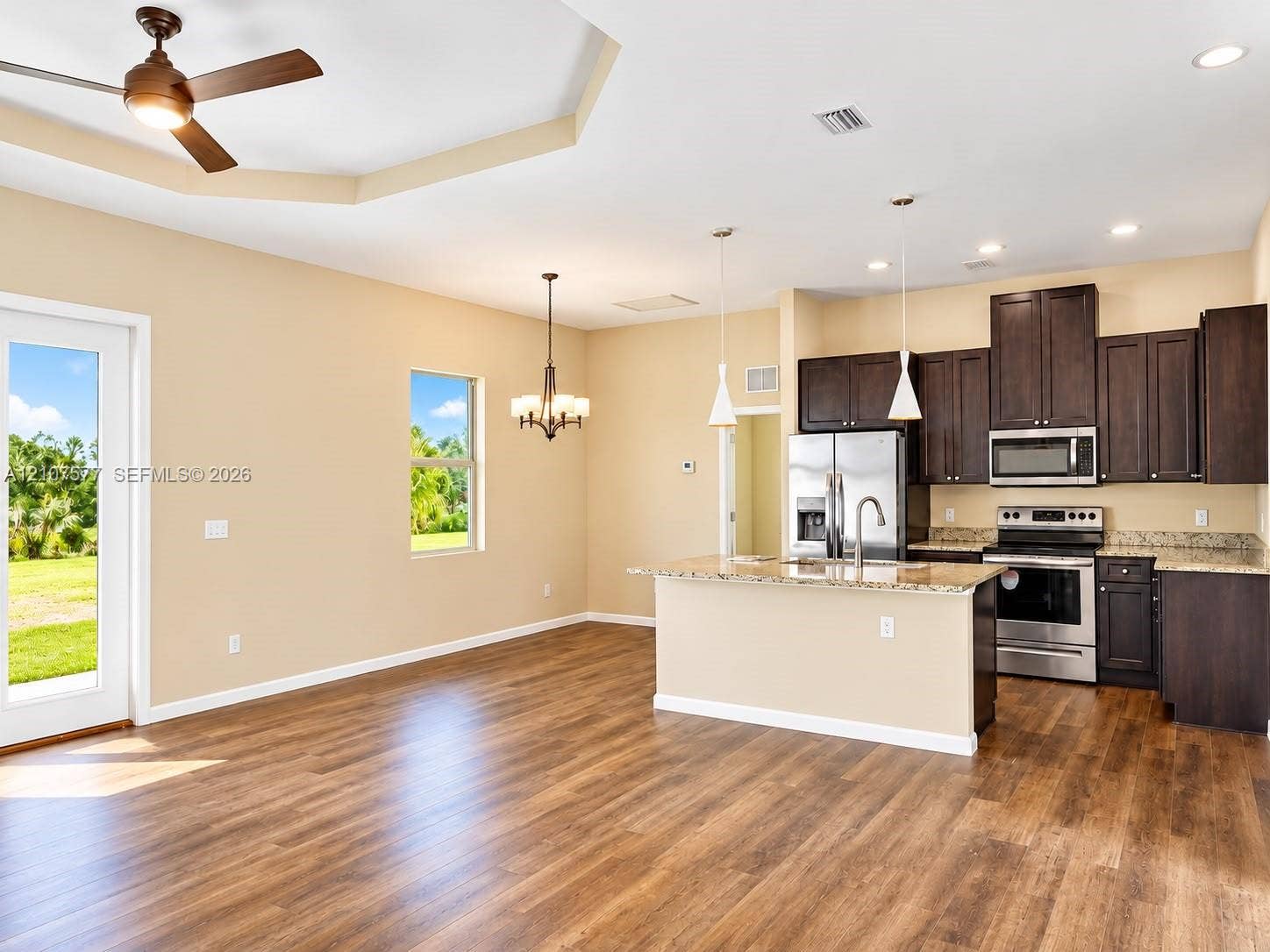 Kitchen featuring dark wood cabinetry, stainless steel appliances, a center island with a stone-finish countertop, wood-finish flooring, and a tray ceiling with a recessed ceiling fan