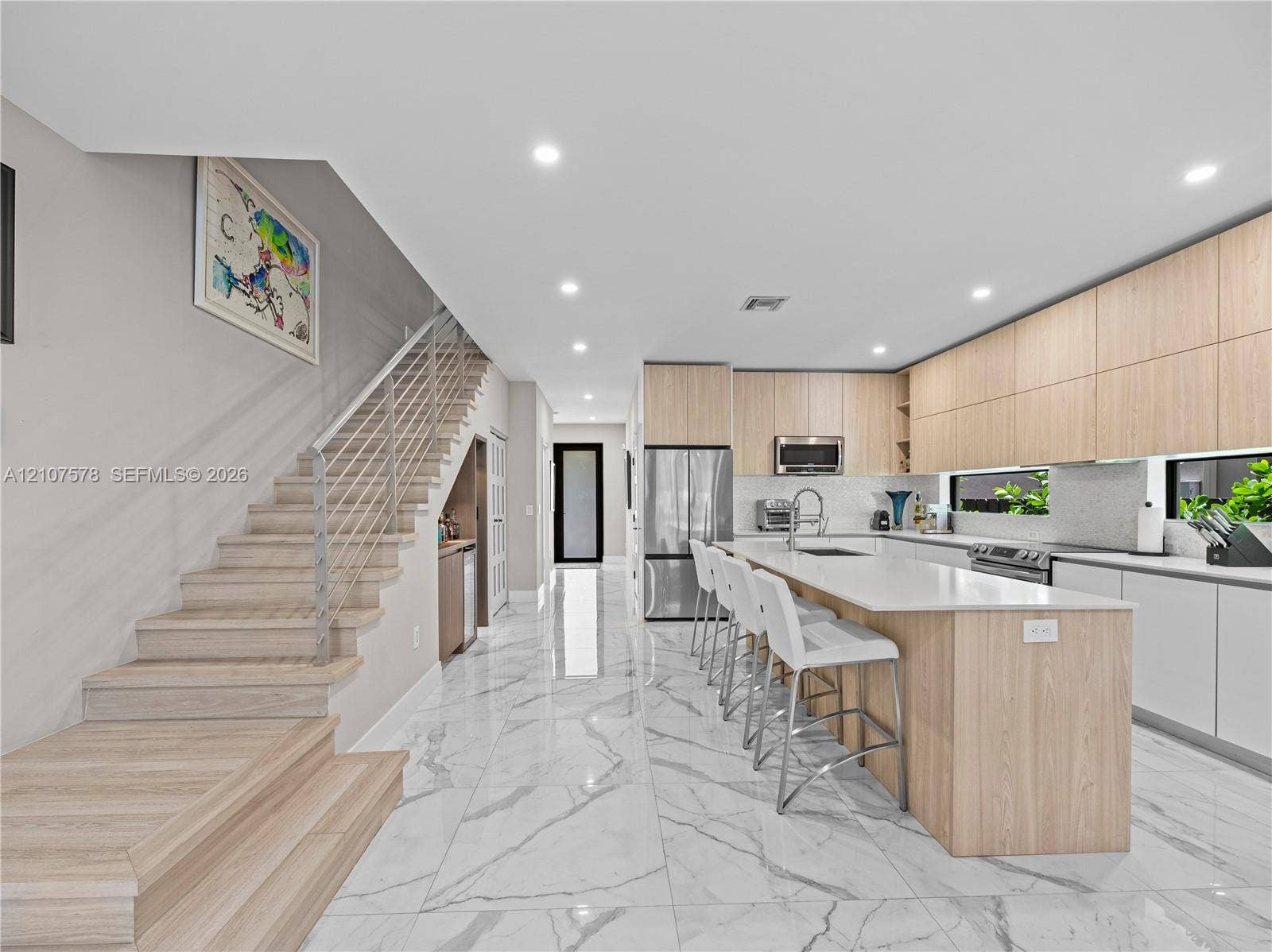307 SW 10th Street, Fort Lauderdale, FL 33315 Modern kitchen featuring light wood-finish cabinetry, a large central island with solid-surface countertops, polished stone-finish floor tiling, and a custom staircase with a sleek horizontal railing