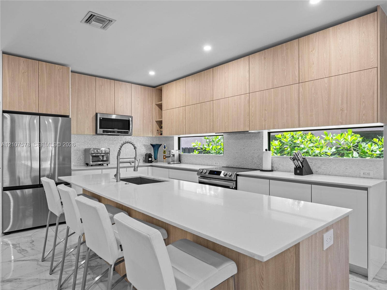 307 SW 10th Street, Fort Lauderdale, FL 33315 Contemporary kitchen featuring light wood-finish cabinetry, white solid-surface countertops, a center island with integrated sink, stainless steel appliances, and ribbon windows