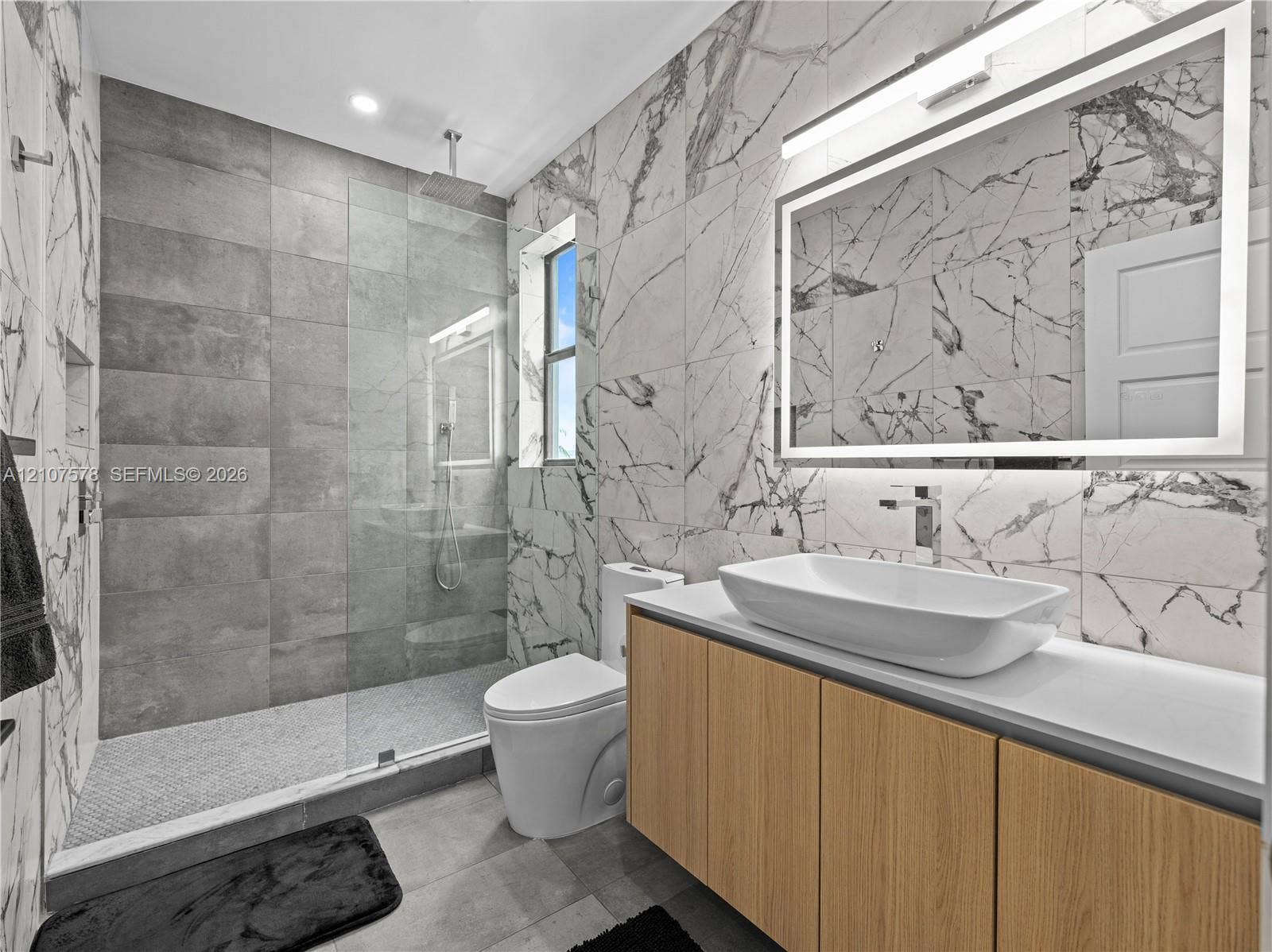 307 SW 10th Street, Fort Lauderdale, FL 33315 Bathroom featuring large-format wall-to-wall stone-style tile, a walk-in glass shower enclosure with a rainfall showerhead, a wood-finish vanity with a rectangular vessel sink, and a large illuminated vanity mirror