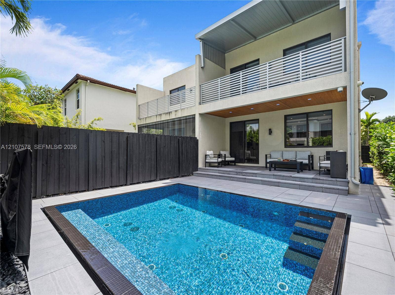 307 SW 10th Street, Fort Lauderdale, FL 33315 Exterior space featuring a rectangular in-ground pool with mosaic tile interior, light-toned stone paver decking, a covered rear patio with wood-finish ceiling, and a horizontal slat privacy fence