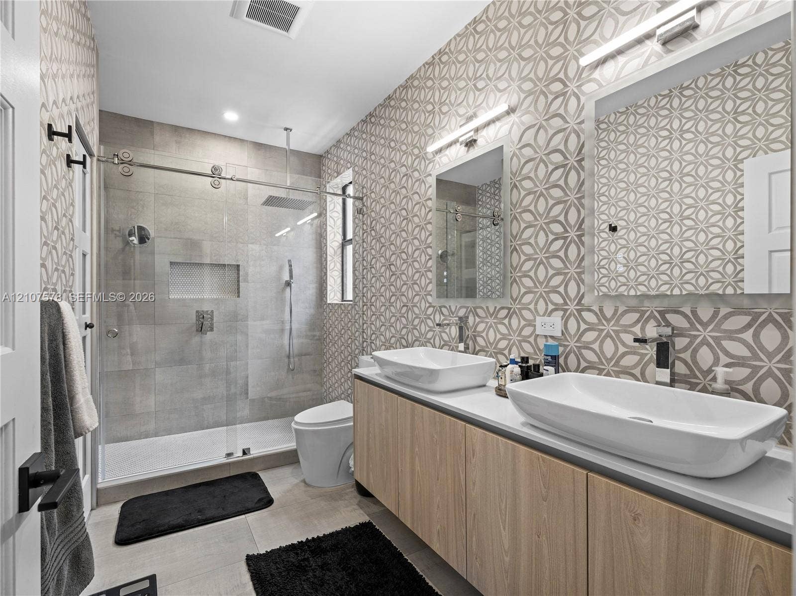 307 SW 10th Street, Fort Lauderdale, FL 33315 Bathroom featuring a dual-sink vanity with light wood-finish cabinetry, patterned wall tile, a frameless glass-enclosed shower with a rainfall showerhead, and wall-mounted faucets