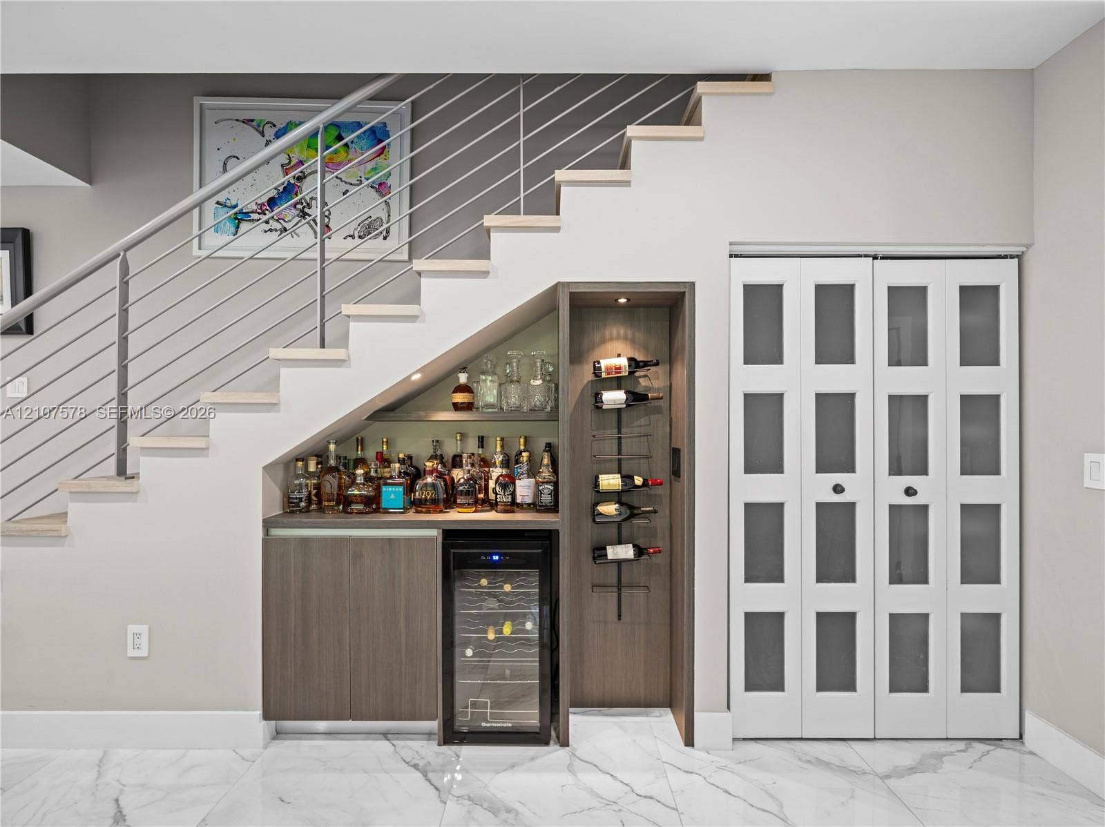 307 SW 10th Street, Fort Lauderdale, FL 33315 Built-in wet bar featuring wood cabinetry, integrated wine refrigerator, vertical wall-mounted wine rack, and recessed accent lighting, positioned beneath a staircase with metal railing and finished with stone-style tile flooring
