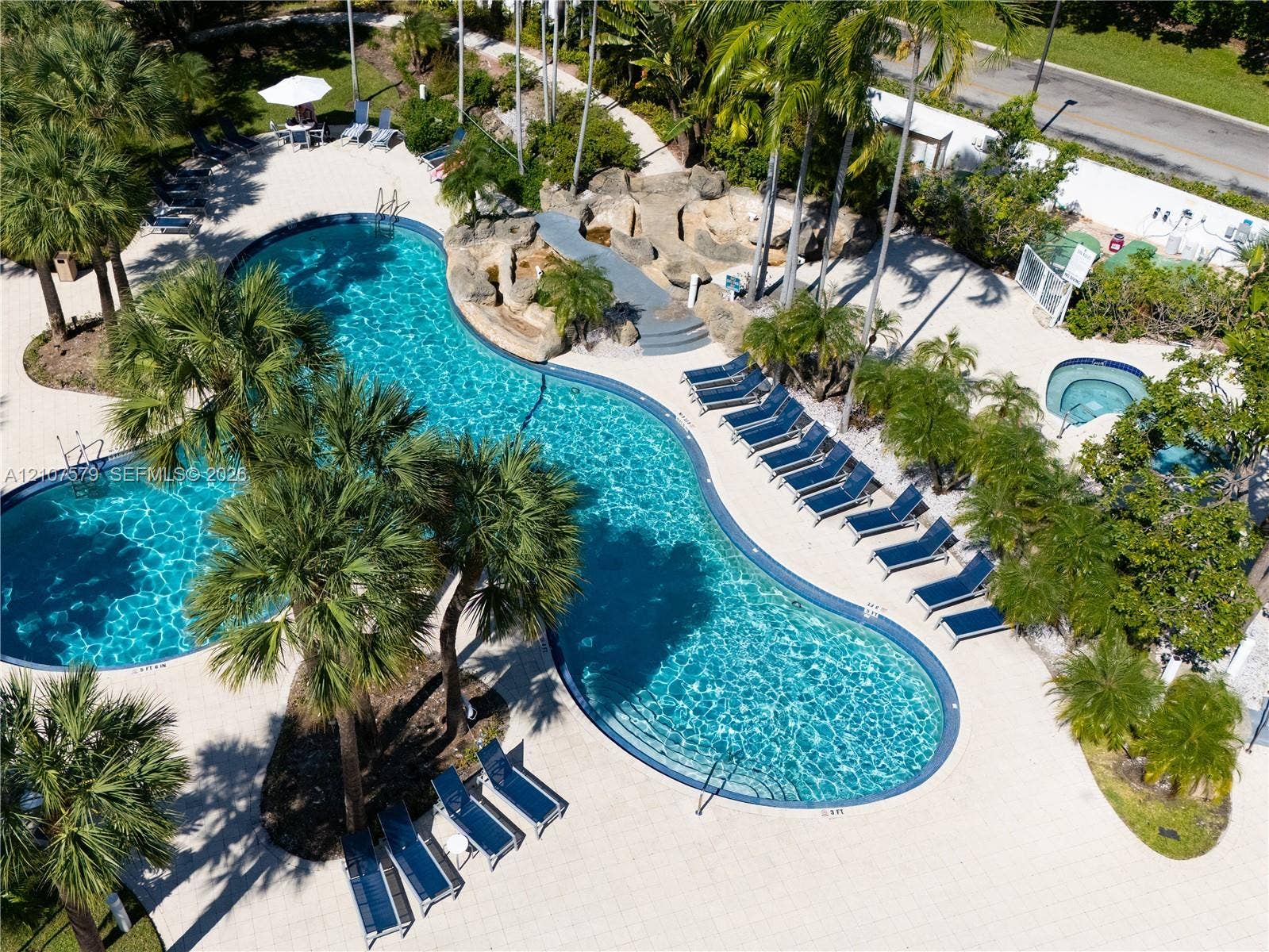 3300 NE 191st Street, #511, Aventura, FL 33180 Curvilinear swimming pool with integrated rock waterfall feature, tiled patio decking, and separate elevated spa, surrounded by mature palm trees