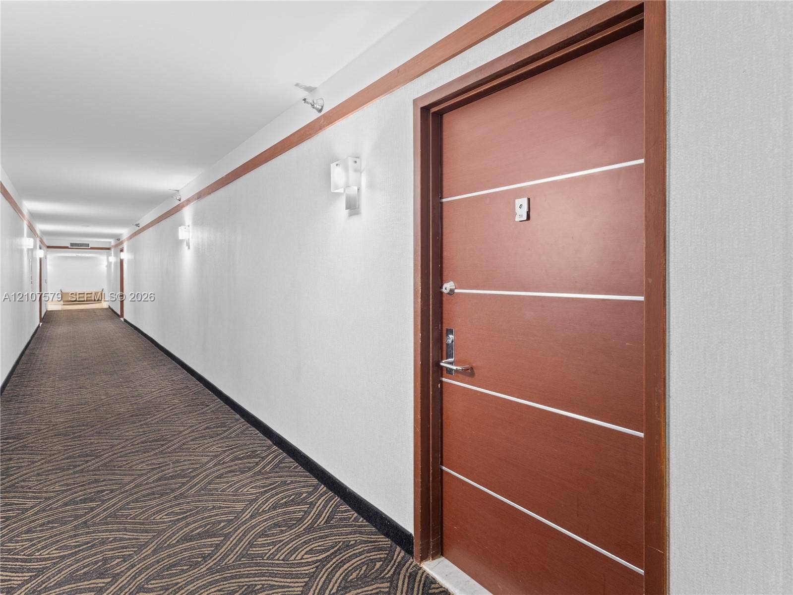 3300 NE 191st Street, #511, Aventura, FL 33180 Interior hallway featuring a wood-finish door with horizontal metal inlay, patterned carpet flooring, wall-mounted sconce lighting, and decorative wood-trim wall accents