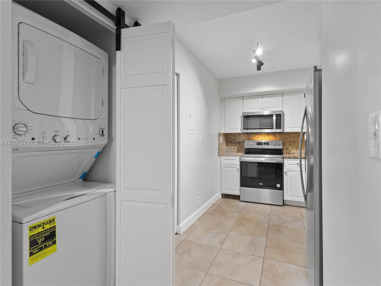 3300 NE 191st Street, #511, Aventura, FL 33180 Laundry closet with stacked washer and dryer behind a sliding door, white shaker-style cabinetry, stainless steel appliances, stone-look tile flooring, and neutral stone-finish backsplash